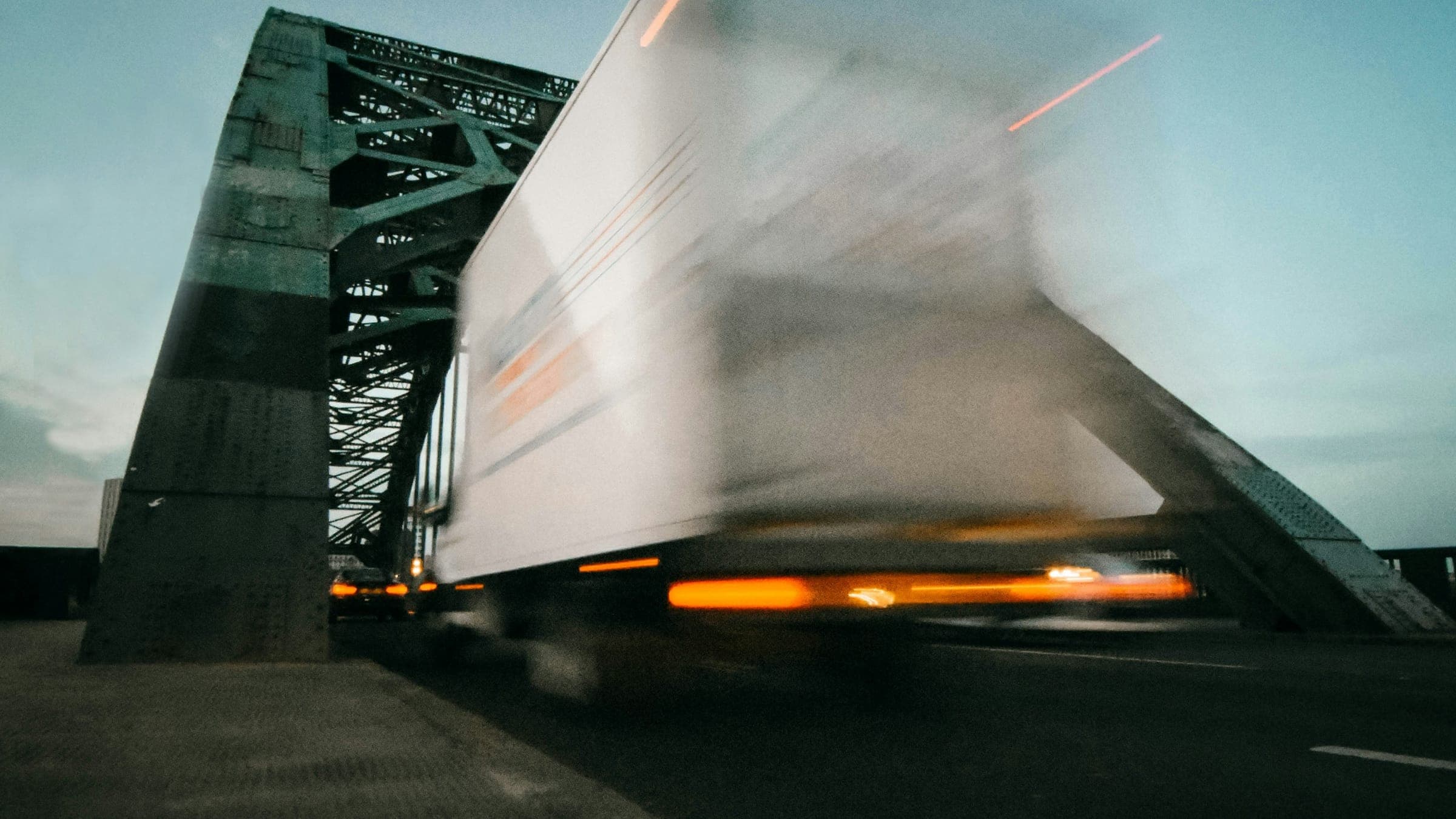 A long-exposure photograph of a tractor-trailer crossing a steel truss bridge at dusk, its lights drawn into streaks.